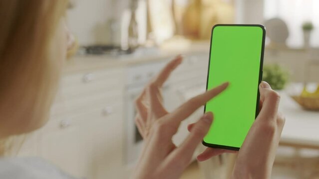 Handheld Camera: Back View of Woman at Kitchen Room Using Phone With Green Mock-up Screen Chroma Key Without Track Points Surfing Internet Watching Content Videos Blogs. Swiping Left Twice.