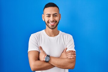 Fototapeta premium Young hispanic man standing over blue background happy face smiling with crossed arms looking at the camera. positive person.