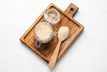 Wooden board with glass jar and spoon of sesame seeds on light background