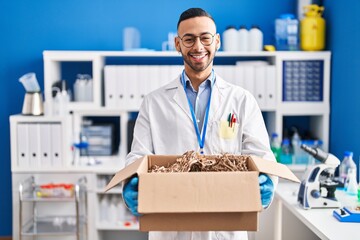 Young hispanic man working at scientist laboratory holding cardboard box celebrating crazy and...
