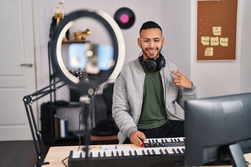 Young hispanic man playing piano at music studio recording himself smiling happy pointing with hand...
