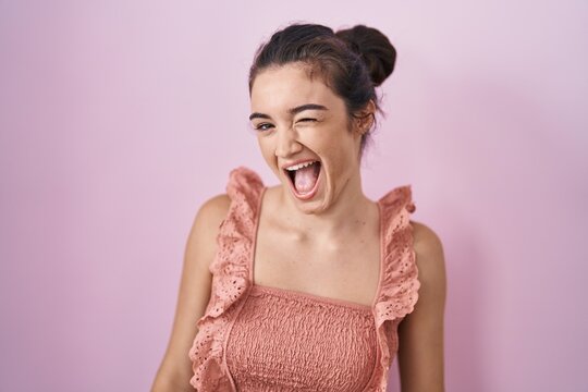 Young teenager girl standing over pink background winking looking at the camera with sexy expression, cheerful and happy face.