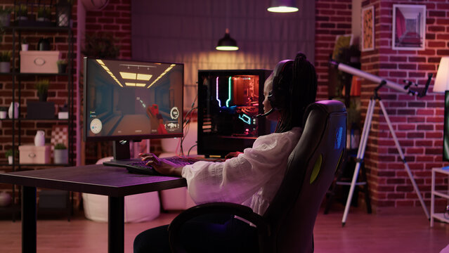 Over Shoulder View Of African American Gamer Girl Using Pc Gaming Setup Playing Multiplayer Streaming Online Action Game In Home Living Room. Woman In First Person Shooter Tournament Talking In