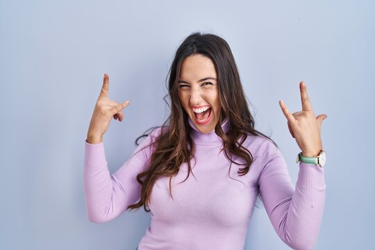 Young brunette woman standing over blue background shouting with crazy expression doing rock symbol with hands up. music star. heavy music concept.