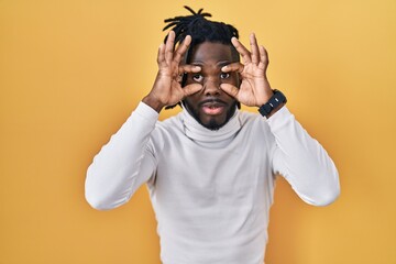 African man with dreadlocks wearing turtleneck sweater over yellow background trying to open eyes with fingers, sleepy and tired for morning fatigue