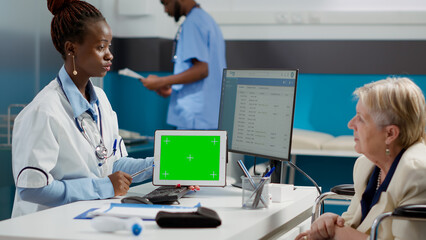 Specialist showing horizontal greenscreen on tablet to paralyzed old woman, looking at isolated chroma key template on display. Medic and patient in wheelchair using blank mockup copyspace.