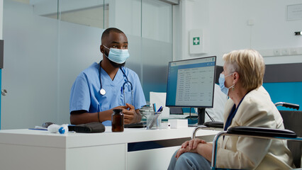 Medical assistant doing consultation with patient wheelchair user, talking about chronic impairment and disability at checkup visit. Nurse consulting paralyzed elderly woman in cabinet.