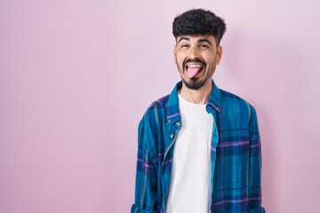 Young hispanic man with beard standing over pink background sticking tongue out happy with funny...