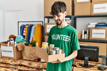Young hispanic man wearing volunteer uniform holding canned food at charity center.