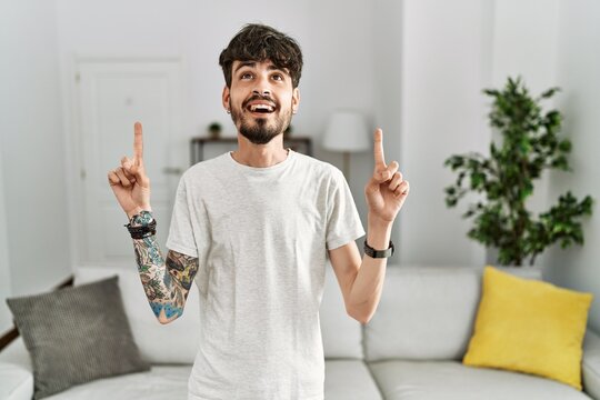 Hispanic man with beard at the living room at home smiling amazed and surprised and pointing up with fingers and raised arms.