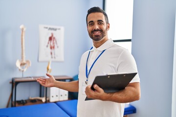 Young hispanic man physiotherapist smiling confident holding clipboard at rehab clinic