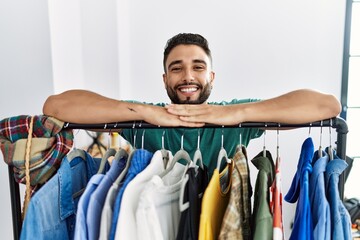 Young arab man customer smiling confident leaning on rack at clothing store