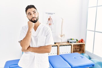 Obraz premium Young handsome man with beard working at pain recovery clinic with hand on chin thinking about question, pensive expression. smiling with thoughtful face. doubt concept.