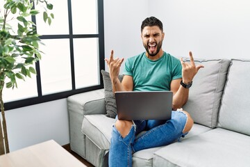 Young handsome man with beard using computer laptop sitting on the sofa at home shouting with crazy...