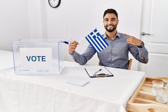 Young handsome man with beard at political campaign election holding greece flag pointing finger to one self smiling happy and proud