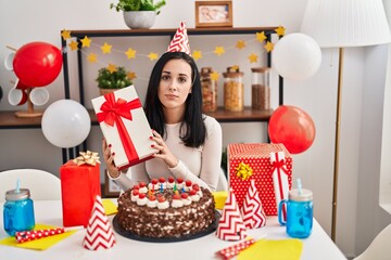 Hispanic woman celebrating birthday with cake holding present relaxed with serious expression on face. simple and natural looking at the camera.