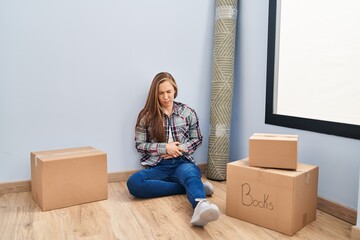 Young blonde woman sitting on the floor moving to a new home with hand on stomach because indigestion, painful illness feeling unwell. ache concept.