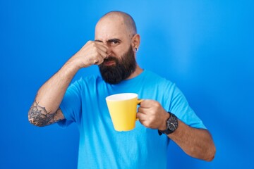 Young hispanic man with beard and tattoos drinking a cup of coffee smelling something stinky and...