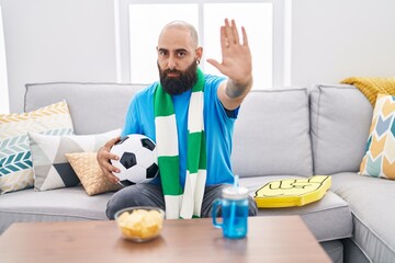 Young hispanic man with beard and tattoos football hooligan holding ball supporting team with open hand doing stop sign with serious and confident expression, defense gesture