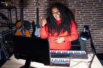 Plus size hispanic woman playing piano at music studio with a big smile on face, pointing with hand...