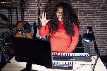Fototapeta premium Plus size hispanic woman playing piano at music studio showing and pointing up with fingers number five while smiling confident and happy.
