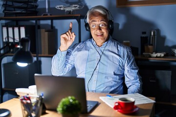 Hispanic senior man wearing call center agent headset at night pointing finger up with successful idea. exited and happy. number one.