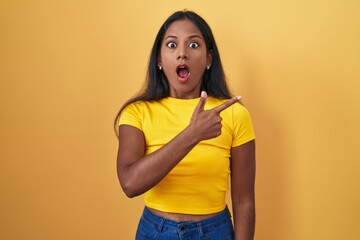 Young indian woman standing over yellow background surprised pointing with finger to the side, open mouth amazed expression.