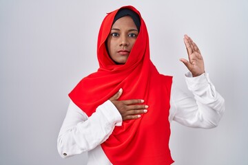 Young arab woman wearing traditional islamic hijab scarf swearing with hand on chest and open palm, making a loyalty promise oath