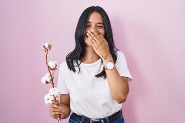 Brunette woman standing over pink background laughing and embarrassed giggle covering mouth with...