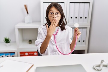 Young hispanic girl wearing doctor coat holding stethoscope covering mouth with hand, shocked and...