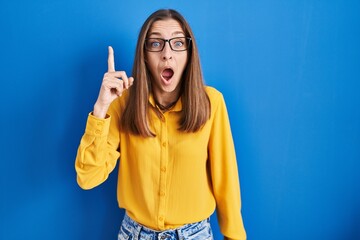 Young woman wearing glasses standing over blue background pointing finger up with successful idea. exited and happy. number one.