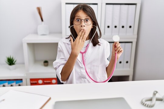 Young hispanic girl wearing doctor coat holding stethoscope covering mouth with hand, shocked and afraid for mistake. surprised expression
