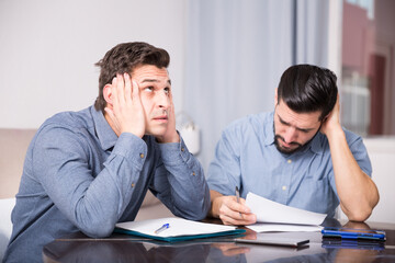Two upset male friends looking worriedly at papers at home table
