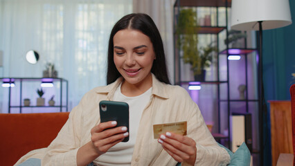 Fototapeta na wymiar Lovely girl using credit bank card and smartphone while transferring money, purchases online shopping, order food delivery at modern home apartment indoors. Young woman in living room lying on couch
