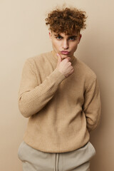 Fototapeta na wymiar a close portrait of a handsome, charming man with curly hair standing in a beige sweater on a beige background, thoughtfully holding his hand on his chin