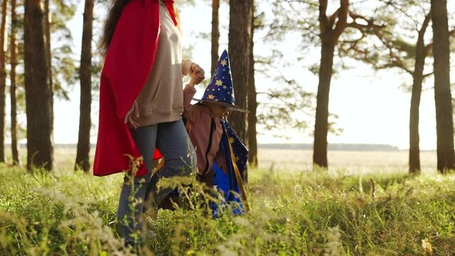 Child plays with his mother in costumes of wizard, playing in park on Halloween. Happy family. Child plays in mantle of magician, mother in red cloak. Childhood dreams, fantasies to become wizard