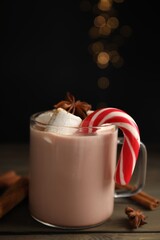 Glass cup of tasty cocoa with marshmallows and Christmas candy cane on wooden table against blurred festive lights