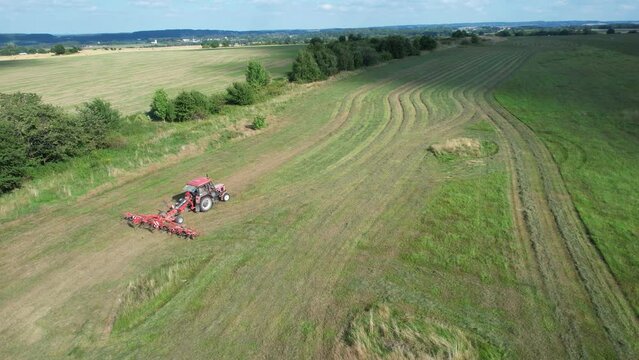 The farmer turns the grass with a hay rake attached to the tractor. Shot from a height.