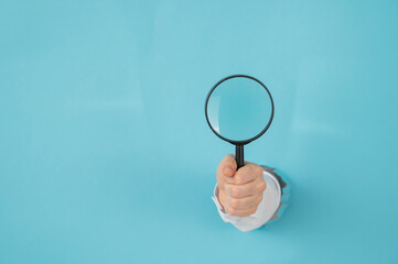 A woman's hand sticking out of a hole in a paper blue background holds a magnifying loupe. Copy space. 