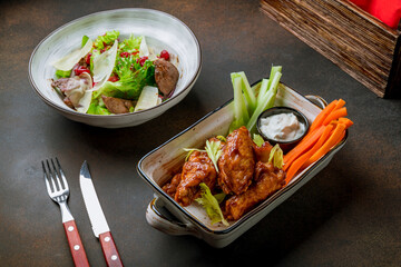 Chicken wings with blue cheese sauce, carrots and celery on plate with chicken liver salad on brown table