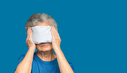 Senior woman using hot compress gel on eyes while standing on a blue background - obrazy, fototapety, plakaty
