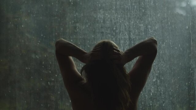 A silhouette of a naked woman playing with her hair and stretching in front of a window with drops of rain. Rear view, close-up, slow motion. Copy space