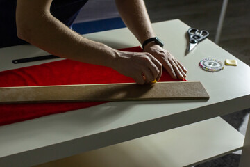 Hands of experienced worker in the handmade industry, performing cutting tasks on red fabric, on a white surface, the beginning of red hat production. Homemade sewing. Working tailor. Christmas hat.