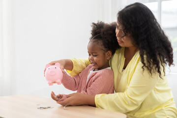 Mother and daughter inserting a coin in a piggy bank. Happy African American kid putting money into the piggybank