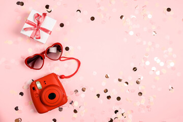 Modern instant camera with sunglasses and gift box on pastel confetti pink background.