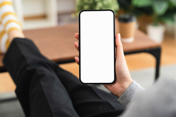 Hands holding smartphone with mockup of blank screen on sofa in living room.