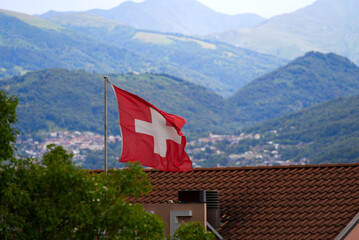 Scenic view over City of Lugano, Canton Ticino, with waving Swiss flag in the foreground on a cloudy summer day. Photo taken July 4th, 2022, Lugano, Switzerland.