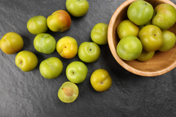 Small mini green fresh juicy plum fruit in wooden bowl on black slate stone board wooden table cut slice half