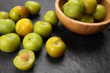 Fototapeta na wymiar Small mini green fresh juicy plum fruit in wooden bowl on black slate stone board wooden table cut slice half