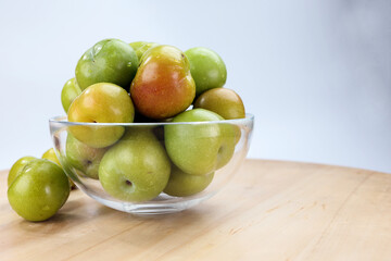 Fototapeta na wymiar Small mini green fresh juicy plum fruit in transparent glass bowl wooden table white background